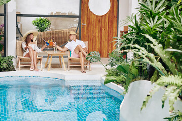 Happy couple relaxing in chaise-lounges by swimming pool and enjoying delicious fruit cocktails