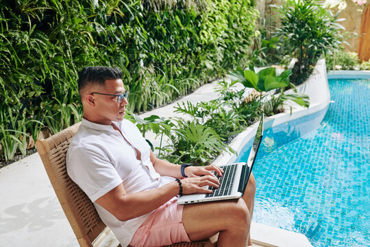 Serious man in glasses sitting by swimming pool and working on laptop, digital nomad concept