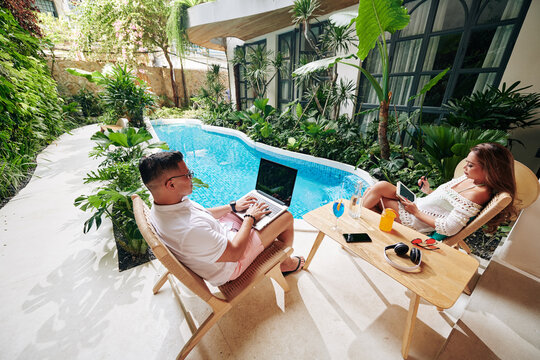 Serious Married Couple Sitting Near Swimming Pool In Backyard, Drinking Cocktails And Working On Gadgets