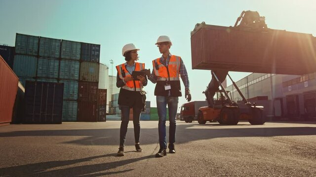 Multiethnic Female Industrial Engineer with Tablet Computer and Male Foreman Worker in Hard Hats and Orange High-Visibility Vests Stand in Container Terminal. Colleagues Talk About Logistics Business.
