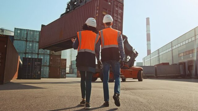 Multiethnic Female Industrial Engineer with Tablet Computer and Male Foreman Worker in Hard Hats and Orange High-Visibility Vests Walk in Container Terminal. Colleagues Talk About Logistics Business.