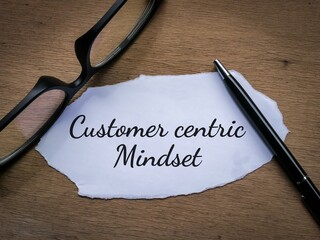 Selective focus top view eye glasses and pen with a piece of paper written Customer centric mindset on wooden background.