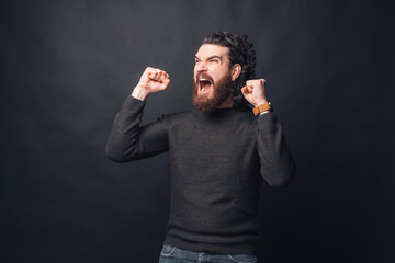 A furious young bearded man is screaming out loud near a black wall.