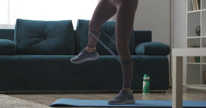 home fitness, woman is training legs with sporty elastic band, tensing muscles, closeup view in living room