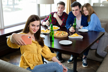 Group or company of young people - friends drink beer, eat pizza, talk and laugh and shoot selfie on the smartphone's camera on the background of the bar