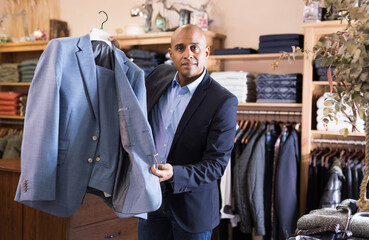 Positive man shop assistant offering suit to customer in menswear boutique