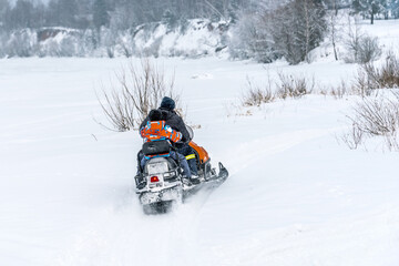 Father rides his son on a snowmobile along the bank of a frozen river