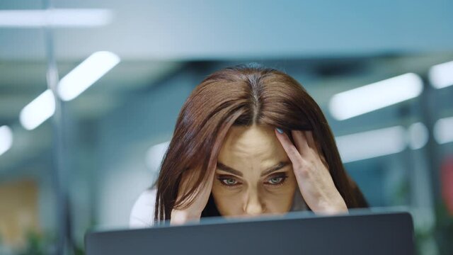Woman with long hair and makeup wearing white shirt sitting at laptop, raising hands and touching hair in despair while receiving message with bad news. Stressed businesswoman in office