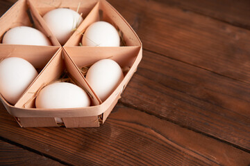 White chicken eggs lie in round wooden basket which stands on a dark wooden table.