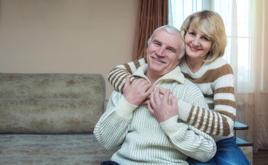 A married couple of 50-60 years old are hugging while sitting on the sofa at home. Looking into the...
