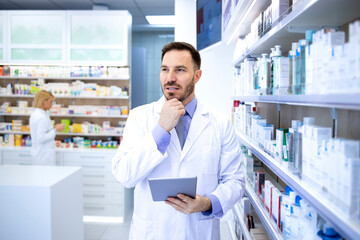 Professional handsome male pharmacist in white coat holding tablet and thinking in pharmacy store...
