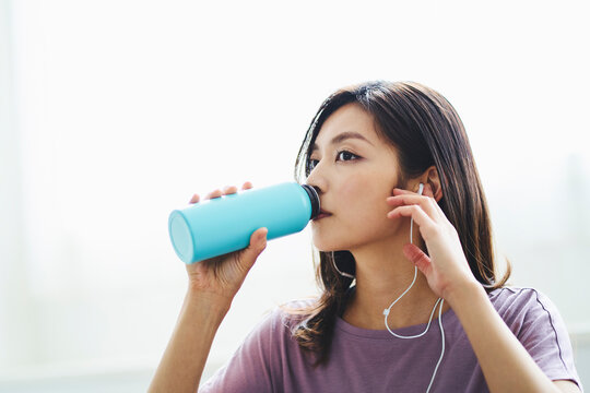 マイボトルで飲み物を飲みながらイヤホンで音楽を聴く30代女性