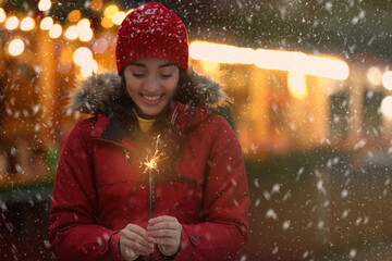 Happy young woman with sparkler at winter fair in evening. Christmas celebration