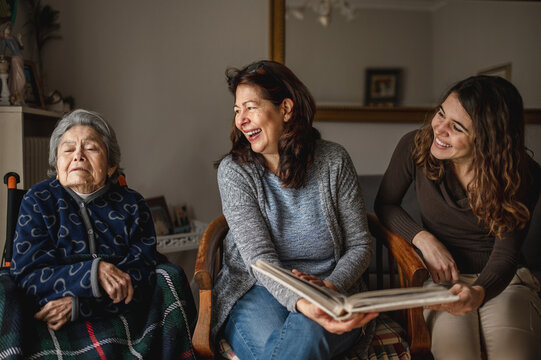 women generation with old sick grandmother sitting in wheelchair and smiling daughter and granddaughter looking a photo album