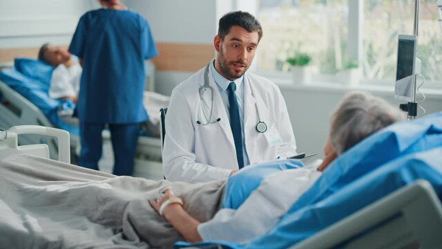Hospital Ward: Friendly Doctor Talks to Beautiful Senior Female Patient Resting in Bed, Explains Test Results, Gives Recovery Advice. Physician Talks to Old Lady Recovering after Successful Surgery