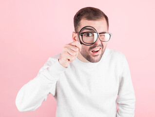 shocked man with glasses with a funny expression in a sweatshirt looks through a magnifying glass on a pink background
