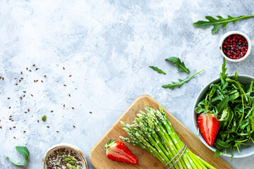 Ingredients for cooking on a gray concrete background. A bunch of fresh green asparagus, strawberries, arugula. Top view. Copy space