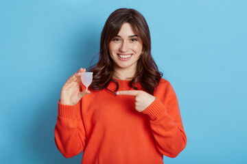 Attractive woman holding menstrual cup and pointing on it with her fore finger, standing against...