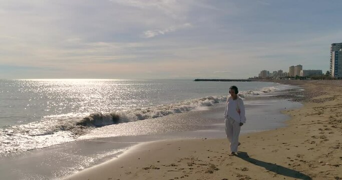 Mujer madura vestida de blanco paseando por orilla de playa y escuchando m&uacute;sica con auriculares