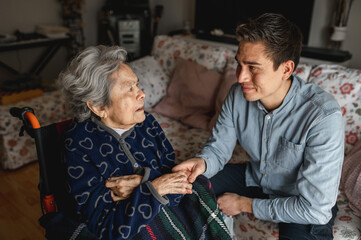 Young man sitting next to an old sick aged woman in wheelchair taking her hands while talking and smiling..