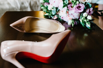 Wedding bouquet of the bride of pink flowers roses and greens, stylish elegant classic lacquered beige shoes and gold rings lying on wooden background. Close up.