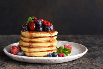 Pancakes with berries and maple syrup for breakfast on a dark concrete background.