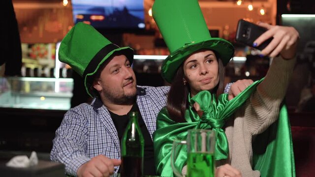 a man and a woman are sitting in a bar celebrating st patrick's day. Big green hats and take a selfie on the phone