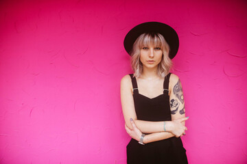 Beautiful young hipster woman with stylish blonde hairstyle, tattoos in black dress and hat posing and looking at the camera isolated on pink background.