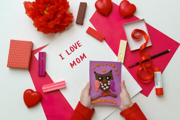 Little blonde girl makes Mother's Day or birthday greeting card and holds a gift in his hands on white table.