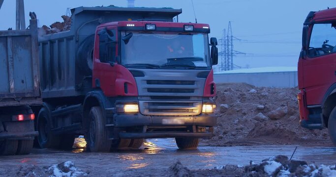Excavation at a construction site, trucks with their headlights on are driving on a muddy road, some are carrying the land, some are waiting for their turn to load 