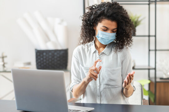 Young African American female student with curly hair in a protective face mask sitting at the desk in a library, spraying hands with sanitizer before touching the laptop, hygiene and safety concept