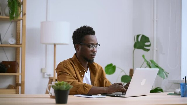 Man Working With Laptop Remotely And Sitting At Table In Home Room Spbas. American African Businessman Looks At Computer Screen And Types, Sits At Desk In Light Interior. Millennial Entrepreneur Doing