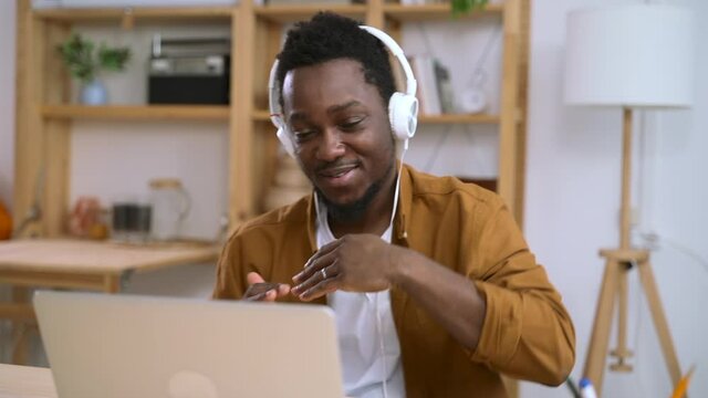 Man having fun and talking in front of laptop screen at table in apartment spbas. Young american guy moves his hands, looks at computer screen and talks with smile while sitting at desk in light room