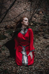 Red-haired woman in a red dress in a historical Celtic costume in the autumn forest