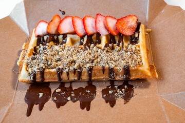 Waffle with strawberry, grated nuts and chocolate sauce in food paper box.