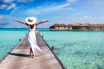 A happy woman in white summer dress and with raised arms walks over a wooden pier in the Maldives islands and enjoys her tropical holidays - obrazy, fototapety, plakaty