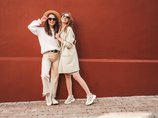Two young beautiful smiling hipster female in trendy white sweater and coat. Sexy carefree women posing near red wall in the street in hat. Positive models having fun, going crazy in sunglasses