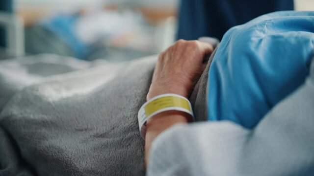 Hospital Ward Bed: Recovering Old Patient Lying in the Hospital Bed Sleeping, Her Fragile Hands Resting on a Blanket, on it Information Wristband. Focus on the Hand. Deeply Emotional Cinematic Shot