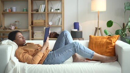 Woman lie on sofa and read book in living room Spbd. African mixed race female student get education, study literature. Concept relax, rest, literacy. Concentrated person at home - Powered by Adobe