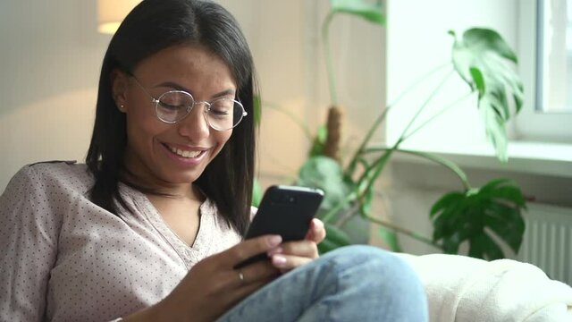 African woman in glasses with smile holds phone in hand background of window Spbd. Portrait of beautiful female model with black skin who is typing text on smartphone. Face of happy person in blouse