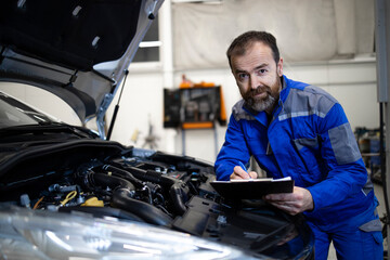 Professional middle aged caucasian car mechanic with checklist standing by vehicle engine area with hood open detecting malfunction.