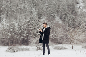 Young man hiker is using smartphone on the snowy mountain. High snowy slopes of mountains and pine forest in snowy winter day on background.