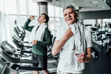 Fototapeta na wymiar The focus is on a senior man in the middle of a modern gym, he wipes himself with a towel and holds a bottle of water in his hands, looking at the camera with a smile. Sports concept