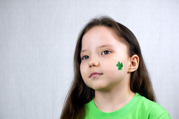 Portrait of cute small girl in green T-shirt with green shamrock leaf on her cheek. Saint Patricks Day holiday