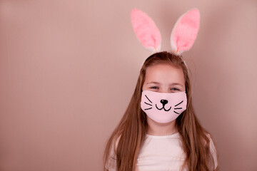 A little Caucasian girl with bunny ears and a medical mask with a painted nose and mustache on her face on a pink background. Easter concept. Coronavirus protected