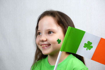 Portrait of smiling endearing small girl in green dress with green shamrock leaf on her cheek and irish flag. Saint Patricks Day celebration