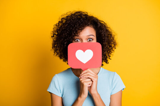 Photo of funky playful dark skin curly girl dressed blue t-shirt holding heart like card cover face isolated yellow color background