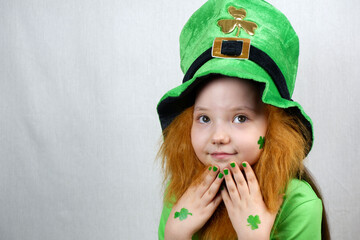 Saint Patricks Day celebration. Nice pensive little girl with decorative red beard, green shamrok leaf on her cheek and leprechaun hat, raised her hands with green nails and clover leaves to her face