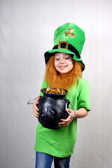 Saint Patricks Day. Funny smiling small girl with decorative red beard, green clover leaf on her cheek and leprechaun hat, dressed in green, looking to cast iron pot, full of leprechaun gold