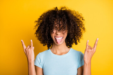 Photo of pretty funny dark skin curly woman dressed blue t-shirt showing hard rock sign tongue isolated yellow color background